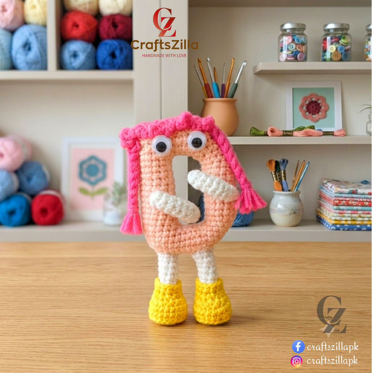 A crocheted letter 'D' amigurumi toy with pink yarn hair, white yarn arms and legs, and yellow yarn feet, standing on a wooden table in a craft room setting. The brand name 'CraftsZilla' is visible in the top left and bottom right corners.