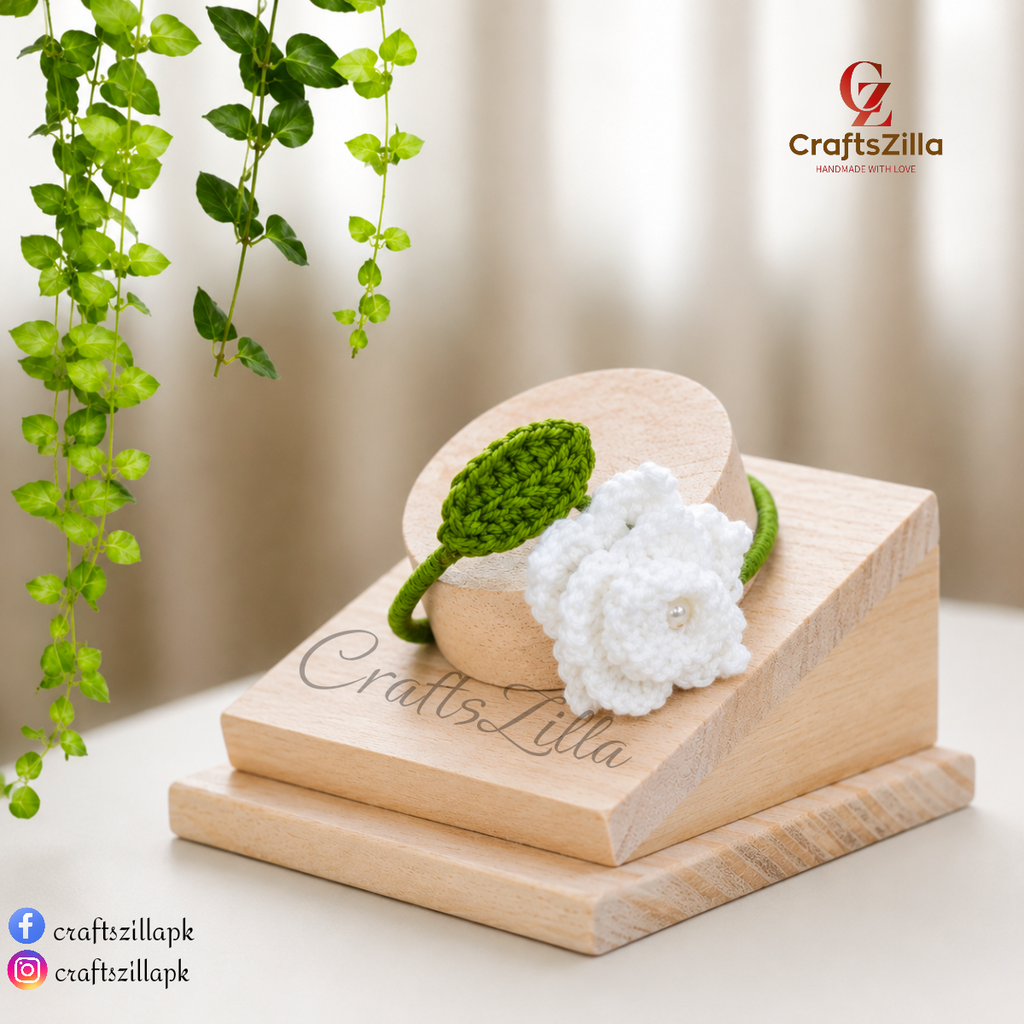A handmade green and white crochet flower bracelet displayed on a wooden stand. The bracelet features a white crochet flower with a small pearl center and a green crochet leaf and bangle.