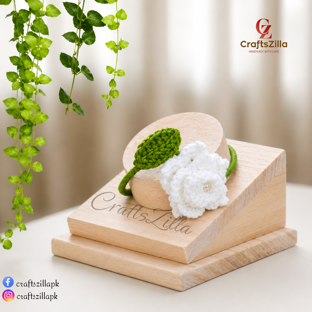A handmade green and white crochet flower bracelet displayed on a wooden stand. The bracelet features a white crochet flower with a small pearl center and a green crochet leaf and bangle.