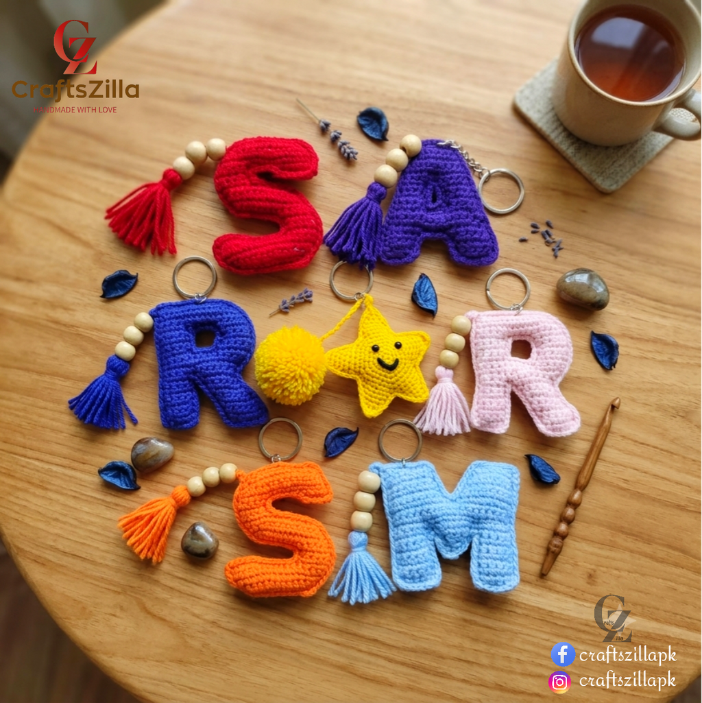 Handmade crochet letter keychains spelling 'ROAR SM' arranged on a wooden table, with decorative beads, stones, a cup of tea, and a wooden spoon. The letters are crocheted in red, purple, blue, yellow, pink, orange, and light blue, with wooden beads and metal keyrings.