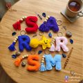 Handmade crochet letter keychains spelling 'ROAR SM' arranged on a wooden table, with decorative beads, stones, a cup of tea, and a wooden spoon. The letters are crocheted in red, purple, blue, yellow, pink, orange, and light blue, with wooden beads and metal keyrings.