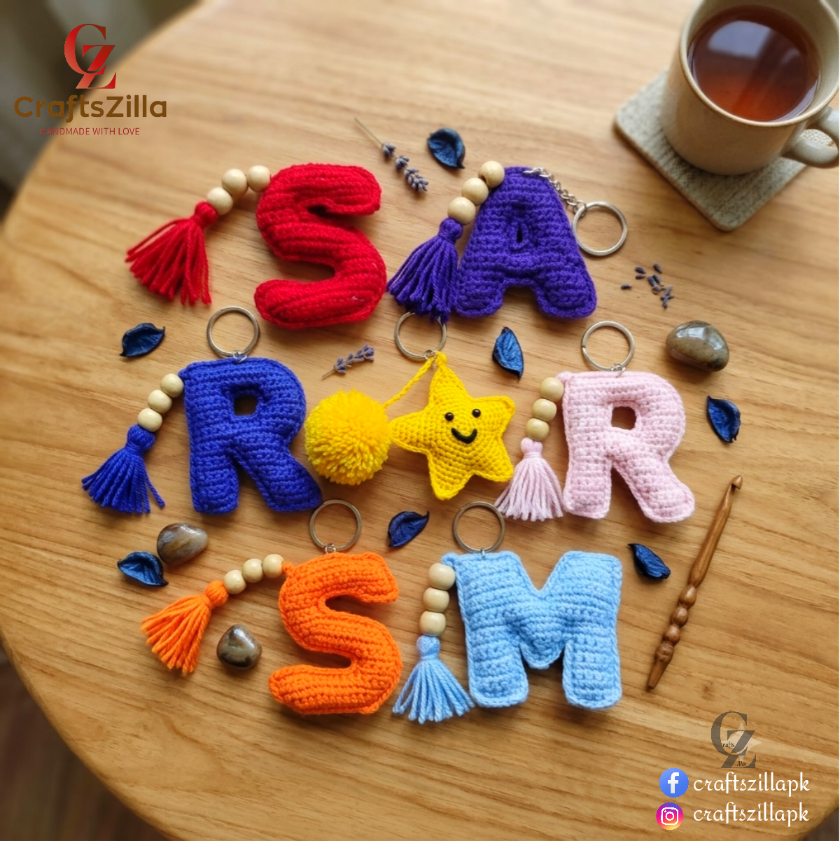 Handmade crochet letter keychains spelling 'ROAR SM' arranged on a wooden table, with decorative beads, stones, a cup of tea, and a wooden spoon. The letters are crocheted in red, purple, blue, yellow, pink, orange, and light blue, with wooden beads and metal keyrings.