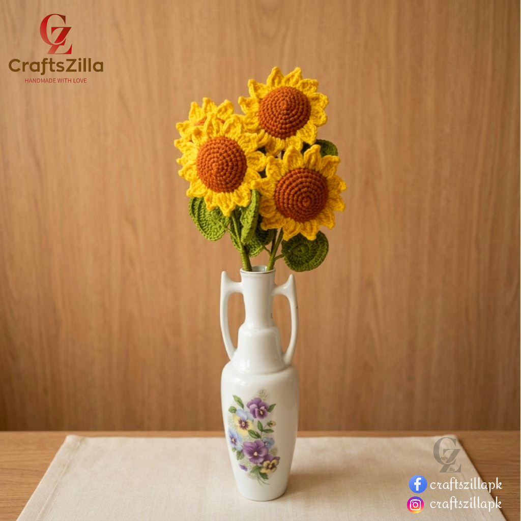Three crocheted sunflower decorations arranged in a white ceramic vase with floral detailing, set on a light fabric surface against a wooden background. Craftzilla branding is visible in the corners.
