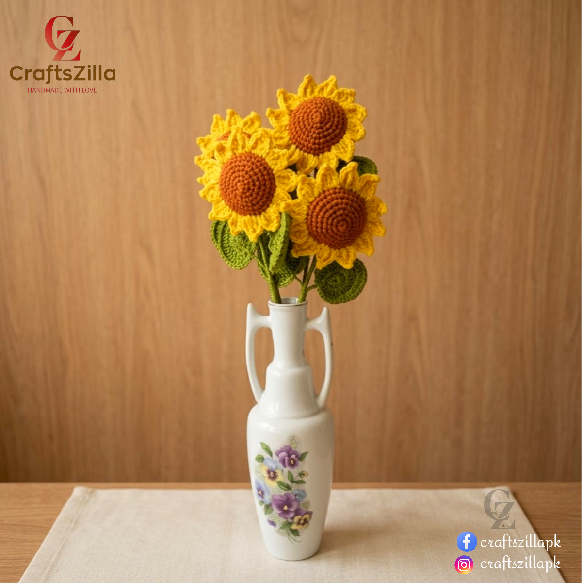 Three crocheted sunflower decorations arranged in a white ceramic vase with floral detailing, set on a light fabric surface against a wooden background. Craftzilla branding is visible in the corners.