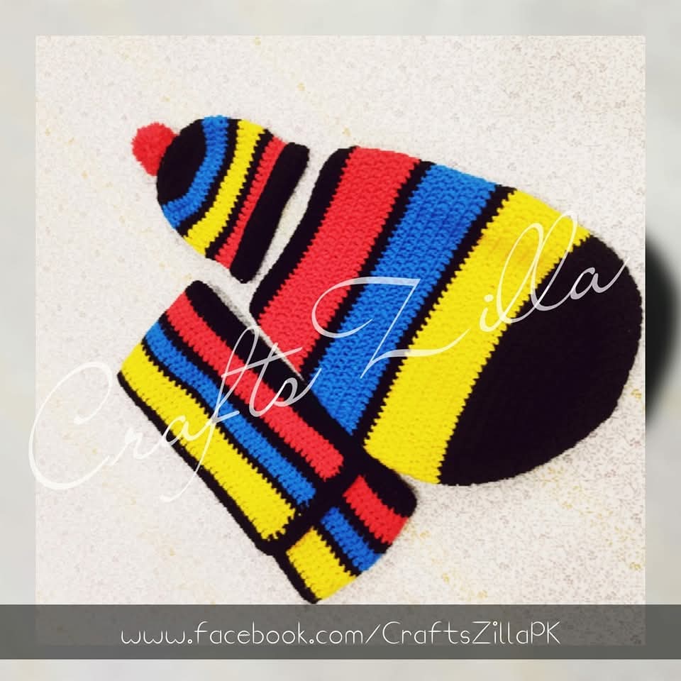 A handmade crocheted knit hat and matching mittens in bold horizontal stripes of red, yellow, blue, black, and white, displayed on a light textured background with a 'Crafts Zilla' watermark.