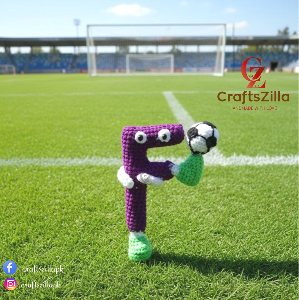 A crocheted letter 'F' toy character with a purple body, green feet, white gloves, and a black-and-white soccer ball, standing on a green soccer field with stadium stands in the background. The CraftsZilla logo is visible in the top right.