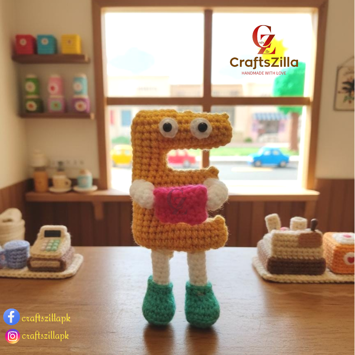 A crocheted letter 'E' toy figure with white yarn eyes, white yarn arms, a pink yarn belly, and green yarn shoes, standing on a wooden table in front of a window. The brand name 'Craftszilla' is visible as an overlay.