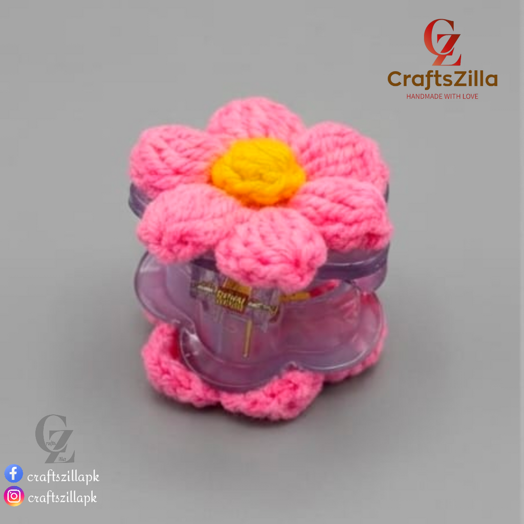 A pink crocheted flower-shaped hair clip with a yellow center, shown on a neutral gray background. The brand logo 'CraftsZilla' is visible in the top right corner.