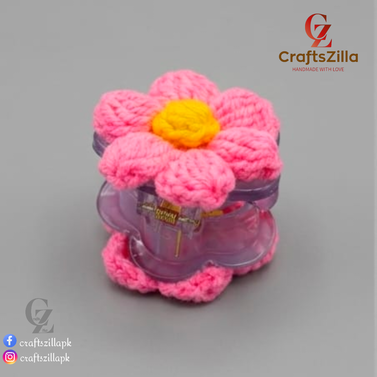 A pink crocheted flower-shaped hair clip with a yellow center, shown on a neutral gray background. The brand logo 'CraftsZilla' is visible in the top right corner.
