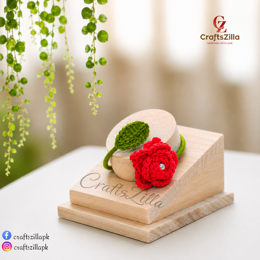 A small handmade red crochet flower bangle bracelet with a green leaf and bangle, displayed on a wooden stand engraved with the brand name CraftsZilla. The CraftsZilla logo is also visible in the top right corner, and social media handles are in the bottom left.