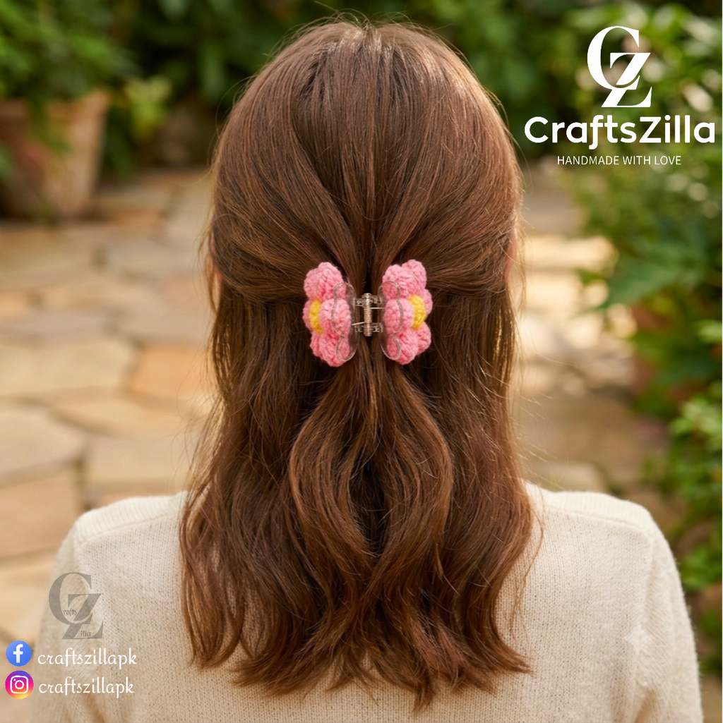 A pink crocheted hair claw clip with a small yellow accent and silver metal clasp, shown on a woman's brown hair from behind in an outdoor garden setting. The 'CraftsZilla' brand logo is visible in the top right and bottom left corners.