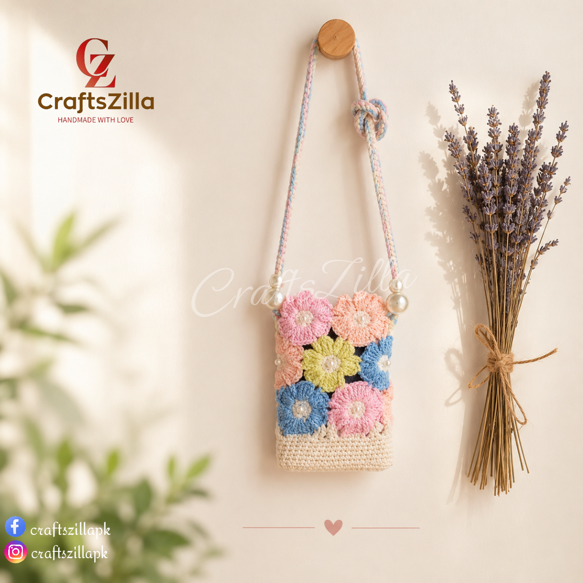 A minimalist lifestyle photograph of a handmade crochet floral crossbody phone pouch hanging from a round wooden wall hook against a warm cream-colored wall. The pouch features pastel pink, peach, yellow, blue, and white granny-square flowers with pearl accents, a cream crocheted base, and a braided pastel shoulder strap finished with pearl beads. A bundle of dried lavender is tied with twine and hangs beside the pouch, while soft sunlight casts gentle shadows across the wall, creating a calm, cozy atmosphe